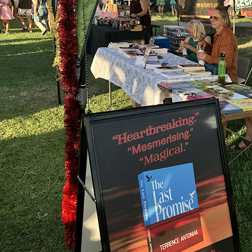 Author market stall at 2023 Bulimba Community Christmas Markets. Terrence and fellow author Tracey Dunn sitting at the table with their books. The Last Promise advertising sandwich board is in the foreground.
