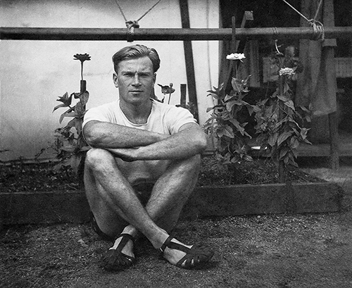 Julius Antoniak sitting cross-legged in front of a flower bed at the door of a wooden-floored canvas temporary accommodation tent at Clyde Migrant Camp, Sydney, Australia in 1949.